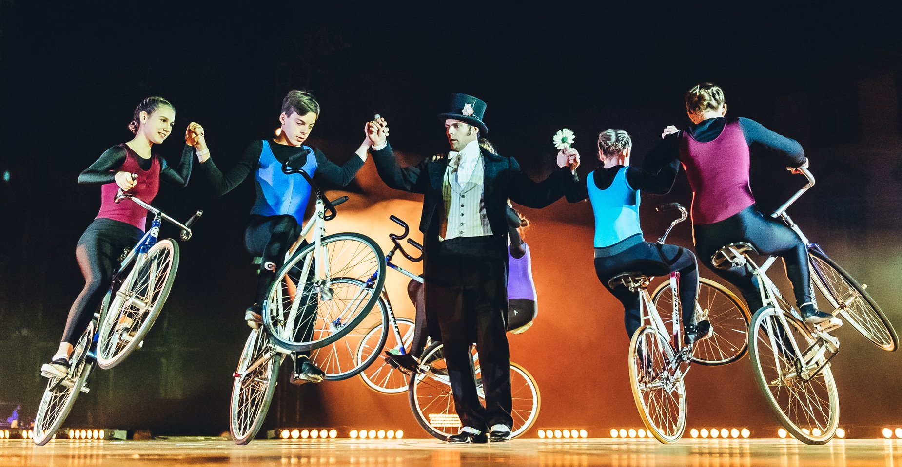 Artistische Fahrradshow bei Nacht – fünf BMX-Fahrer posieren synchron mit einem Performer in der Mitte vor leuchtender Kulisse.