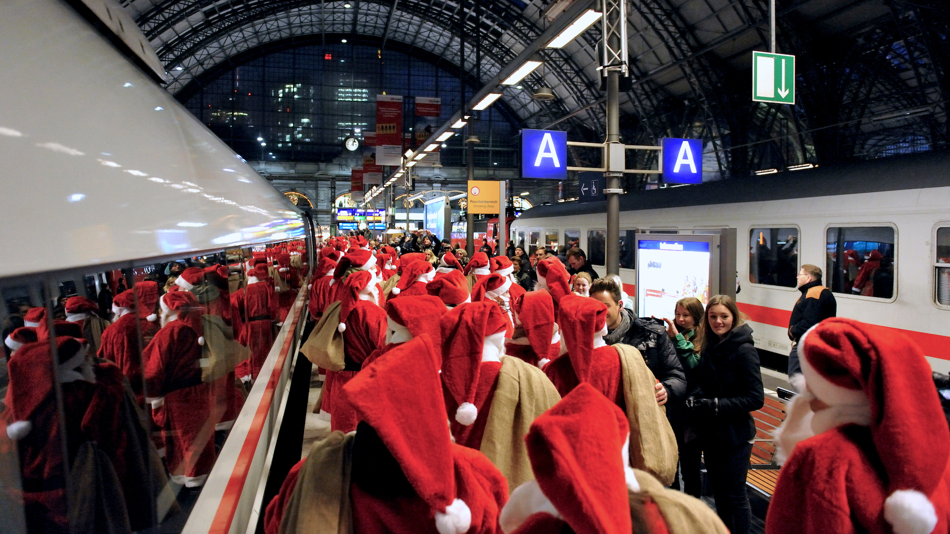 Zahlreiche als Nikolaus verkleidete Menschen warten auf dem Bahnsteig eines großen Bahnhofs.