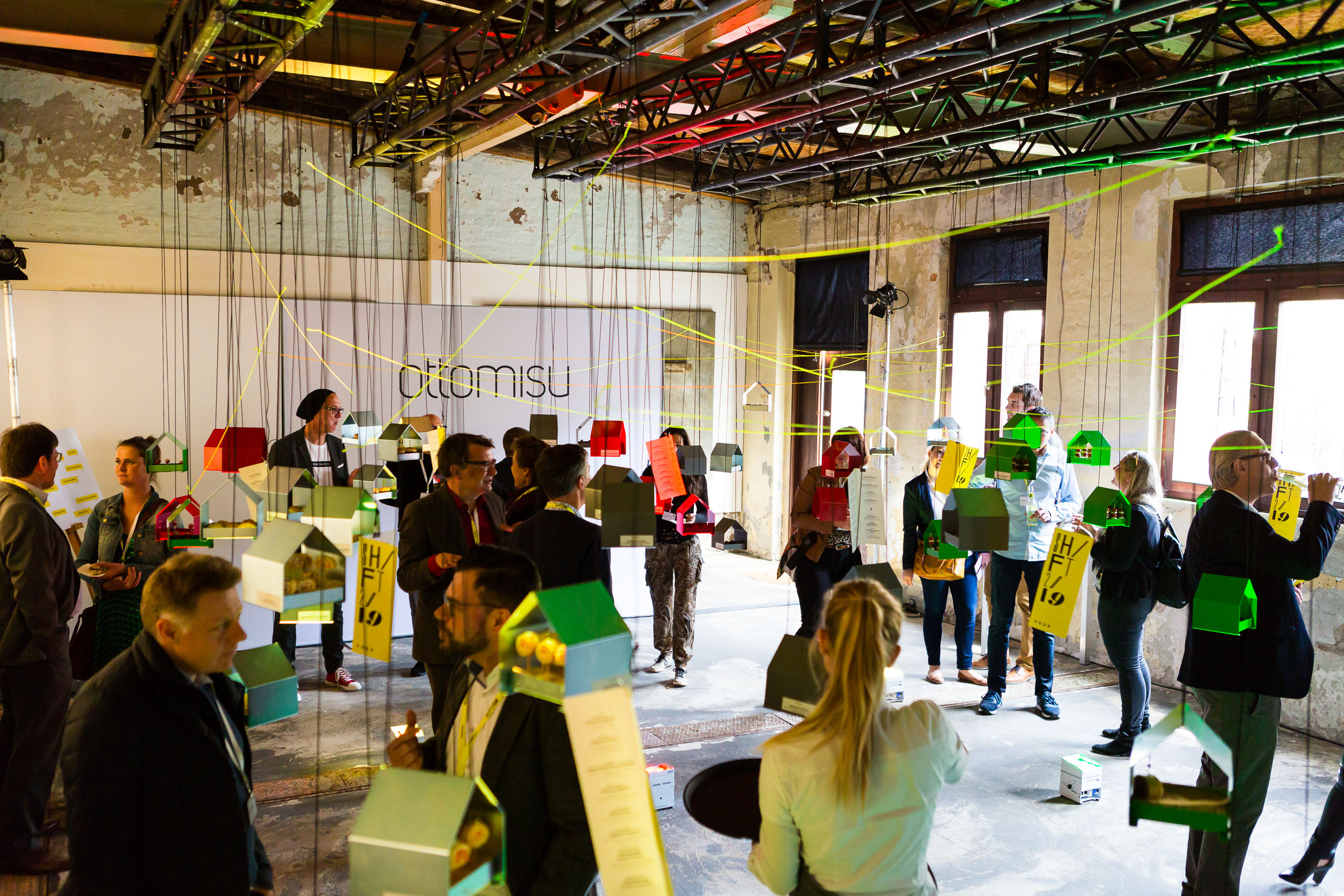 Besucher erkunden einen kreativ gestalteten Ausstellungsraum mit schwebenden Miniaturhäusern in verschiedenen Farben.