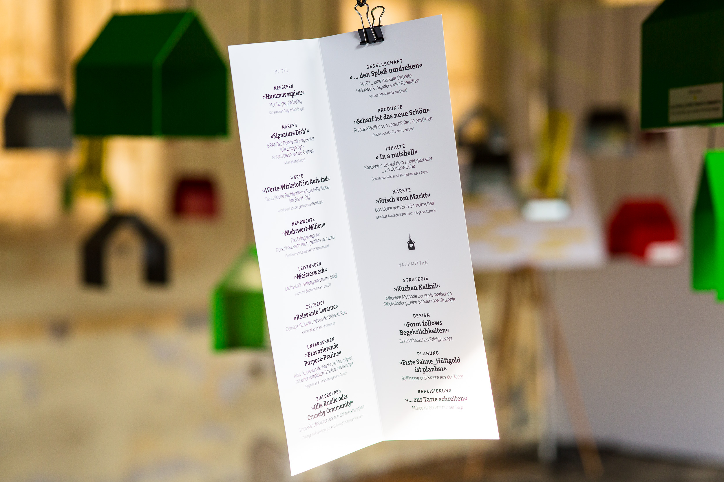 Hängendes Veranstaltungsprogramm mit Logos der Speaker, im Hintergrund ein lichtdurchfluteter Raum mit farbigen Häusern als Dekoration.