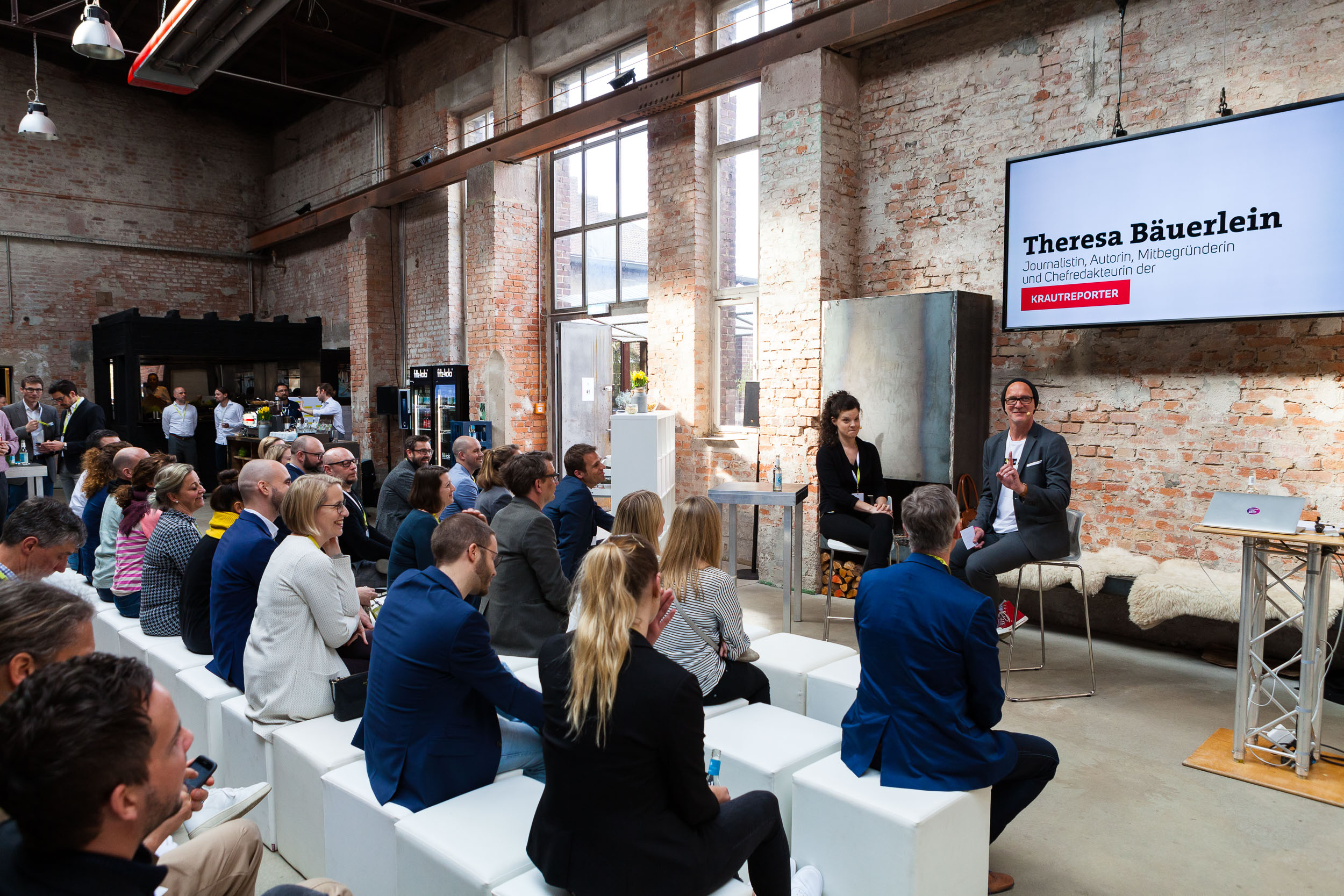 Besucher eine Podiumsdiskussion mit Präsentationsbildschirm in einem lichtdurchfluteten Eventraum.