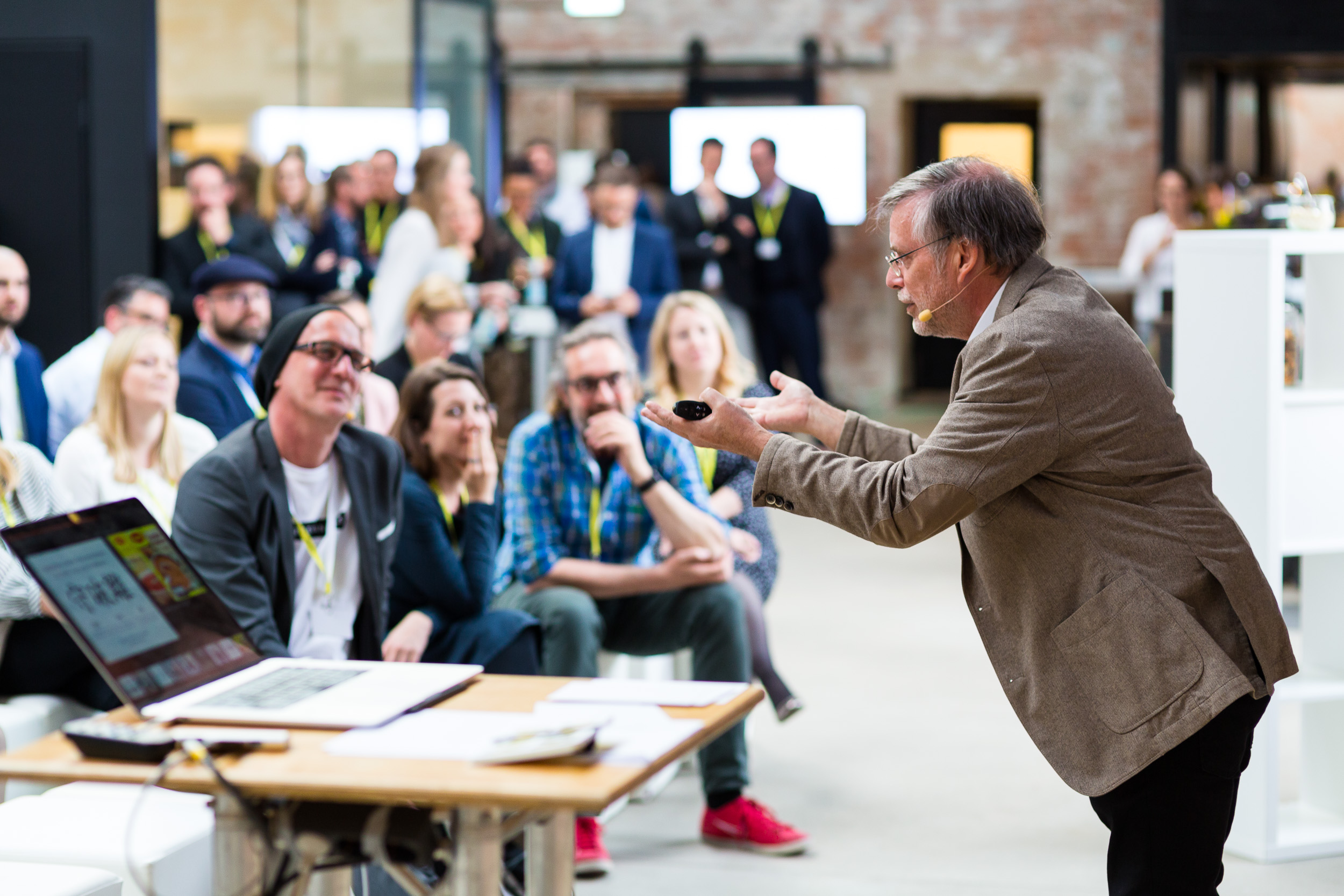 Begeisterter Redner spricht vor aufmerksamem Publikum in einer offenen Industriehalle während eines Events oder Workshops. Ein Laptop und Materialien liegen auf einem Tisch im Vordergrund.
