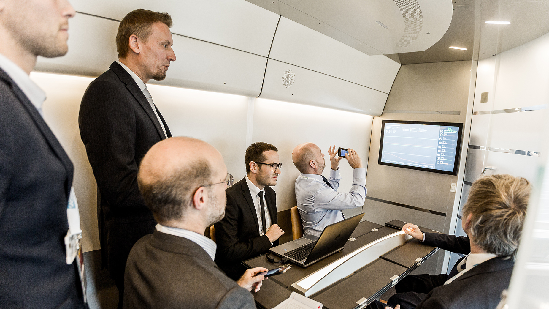 Gruppe in formeller Kleidung bei einer Präsentation im Zug, Blick auf Bildschirm und Laptops.