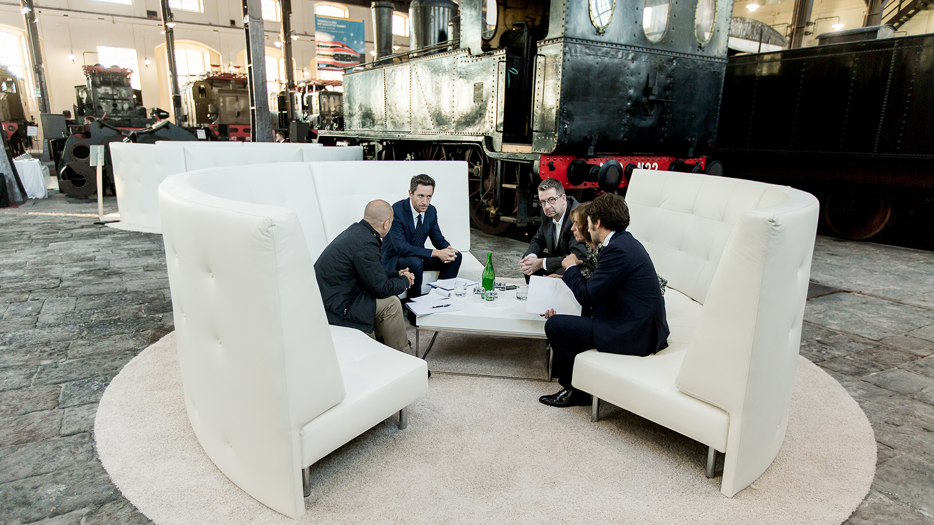 Drei Männer im Gespräch in einer weißen Lounge-Sitzgruppe in offener Eventhalle.