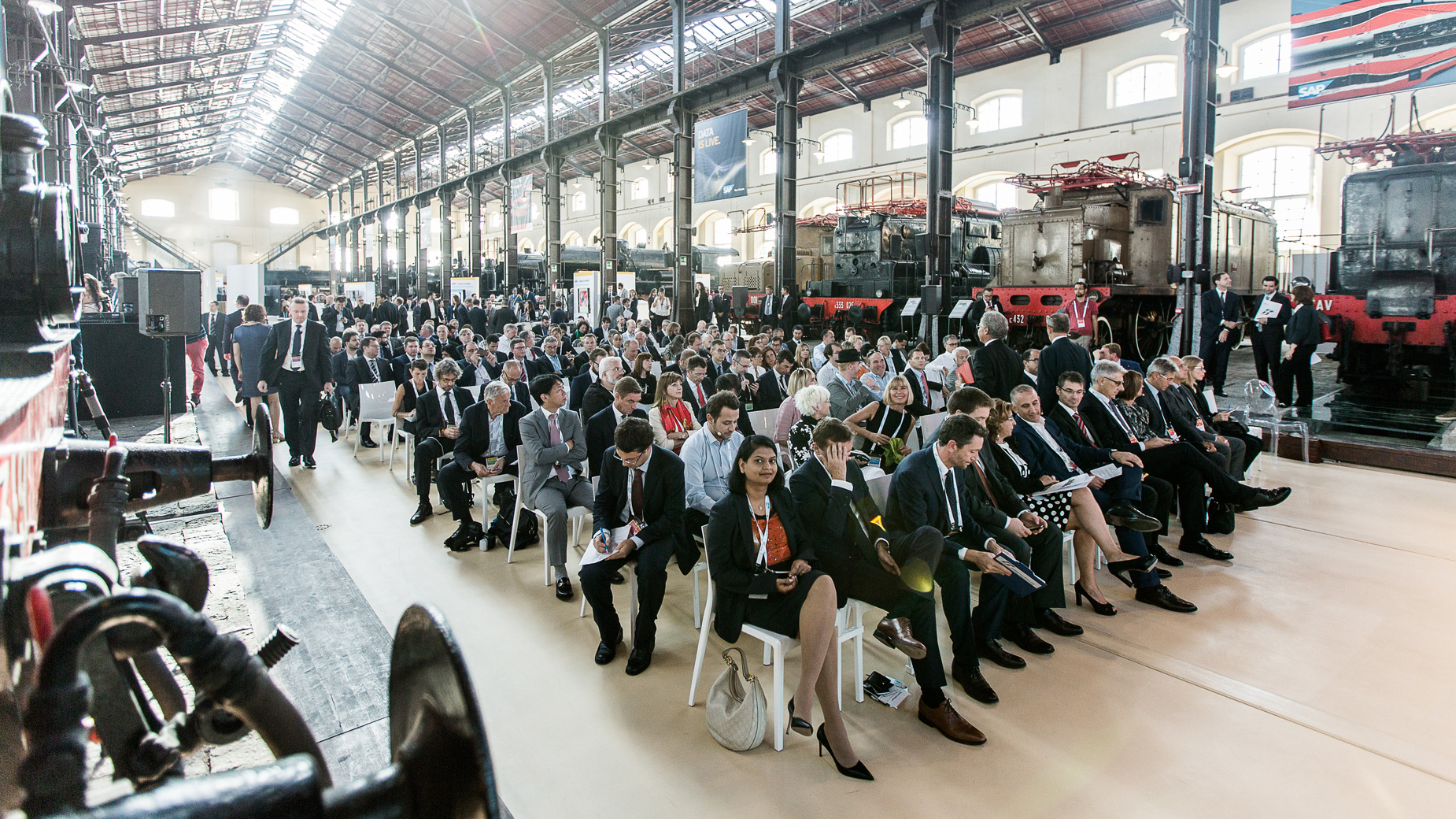 roße Zuhörergruppe auf Stuhlreihen in einer Zughalle, aufmerksam bei einer Präsentation.