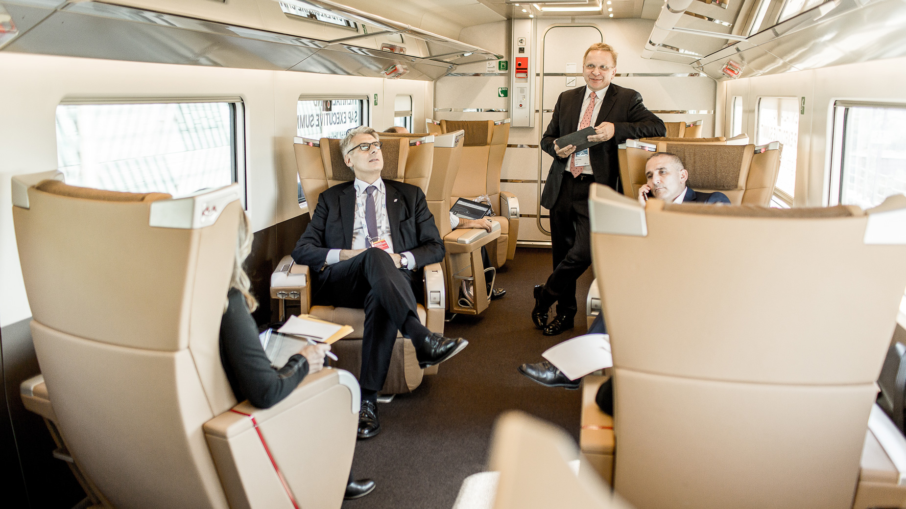 Business-Männer im Austausch in einer komfortablen Zug-Sitzgruppe mit goldfarbenen Sitzen.