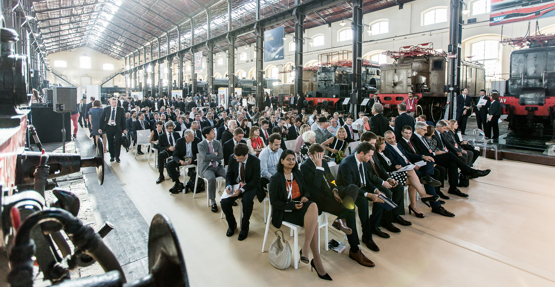Große Zuhörergruppe auf Stuhlreihen in einer Zughalle, aufmerksam bei einer Präsentation.
