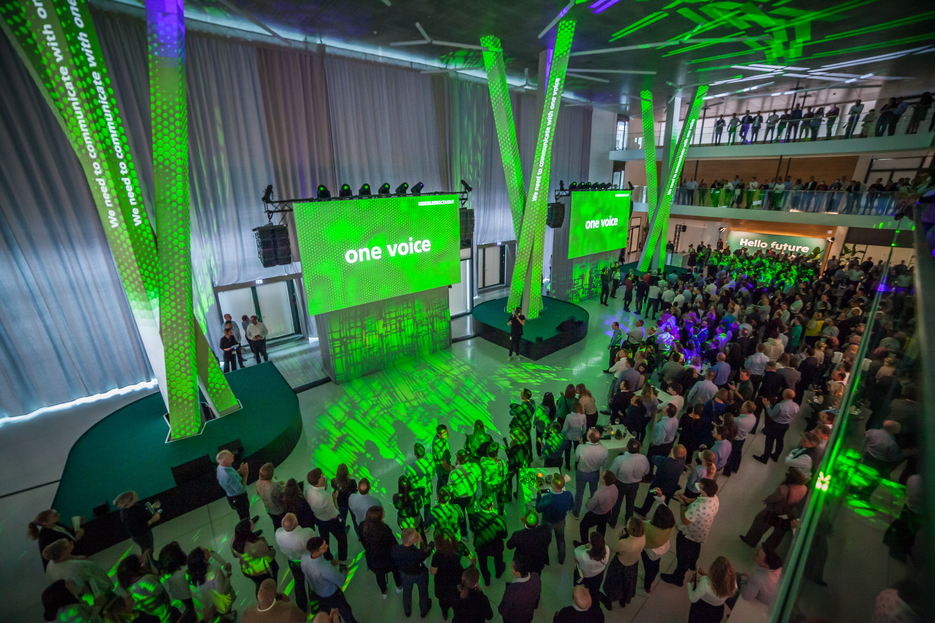 Großes Corporate Event in einem modernen, mehrstöckigen Gebäude mit grüner Lichtinstallation. Auf zwei LED-Wänden steht „one voice“. Zahlreiche Gäste stehen und schauen zur Bühne, während auf der oberen Galerie weitere Personen zusehen.