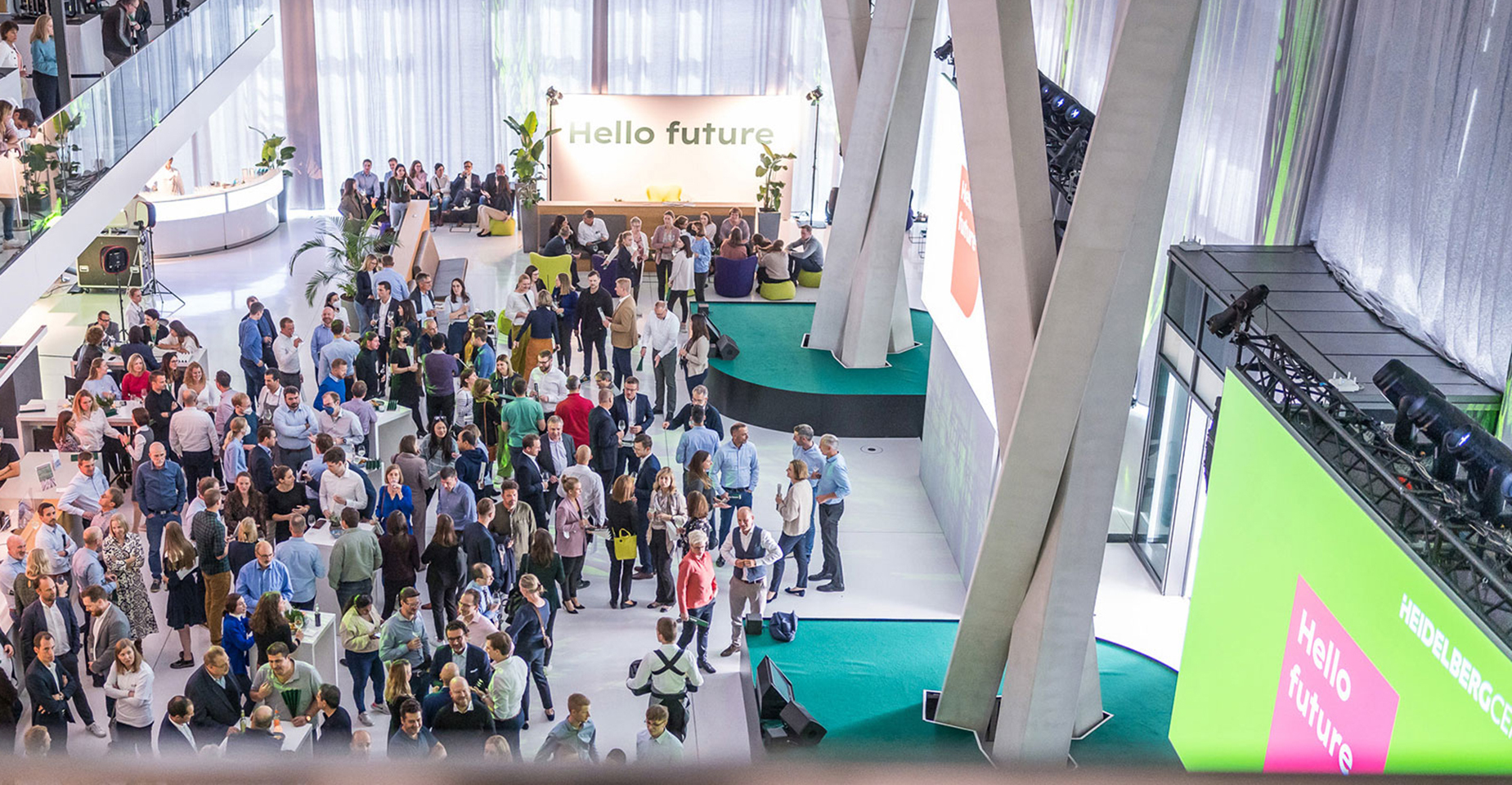 Vogelperspektive auf eine Veranstaltung im Atrium. Besucher:innen stehen in Gruppen zusammen, eine große Leinwand mit grünem Design ist gut sichtbar.