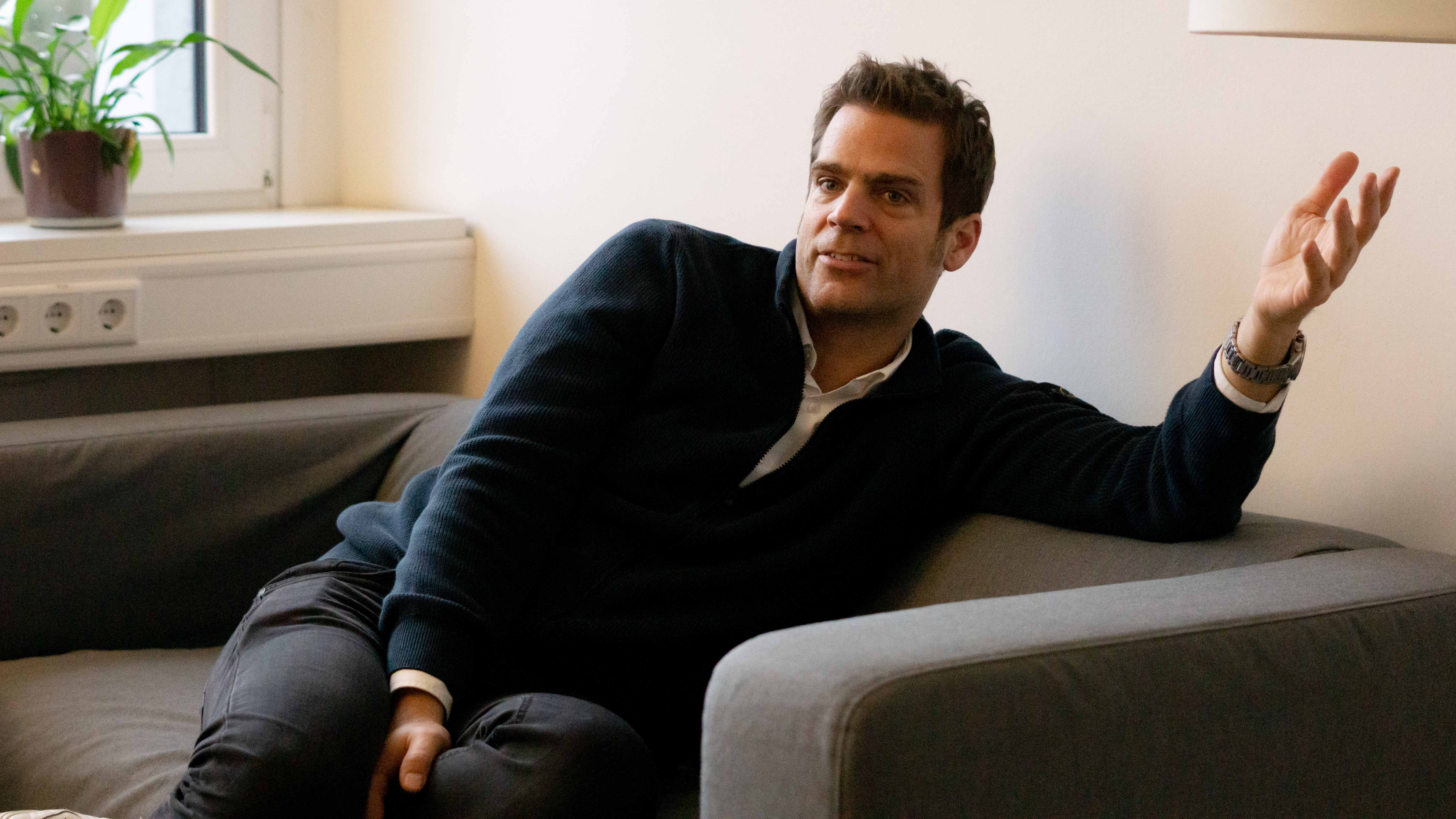 ottomisu CEO Jörn Huber sitzt auf einem grauen Sofa im Büro und führt ein Interview mit Kolleg*innen.