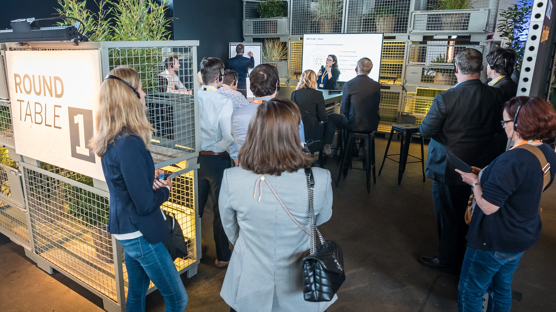 Die Besucher*innen stehen an einem Roundtable mit Gitterkästen, in dem ein Austausch stattfindet.