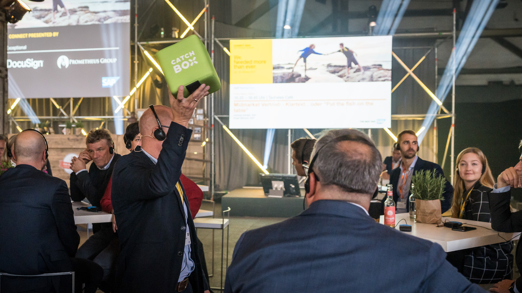 Besucher*innen im Vordergrund, die an den Tischen stehen und sitzen, im Hintergrund befindet sich die Hauptbühne der CONNECT.