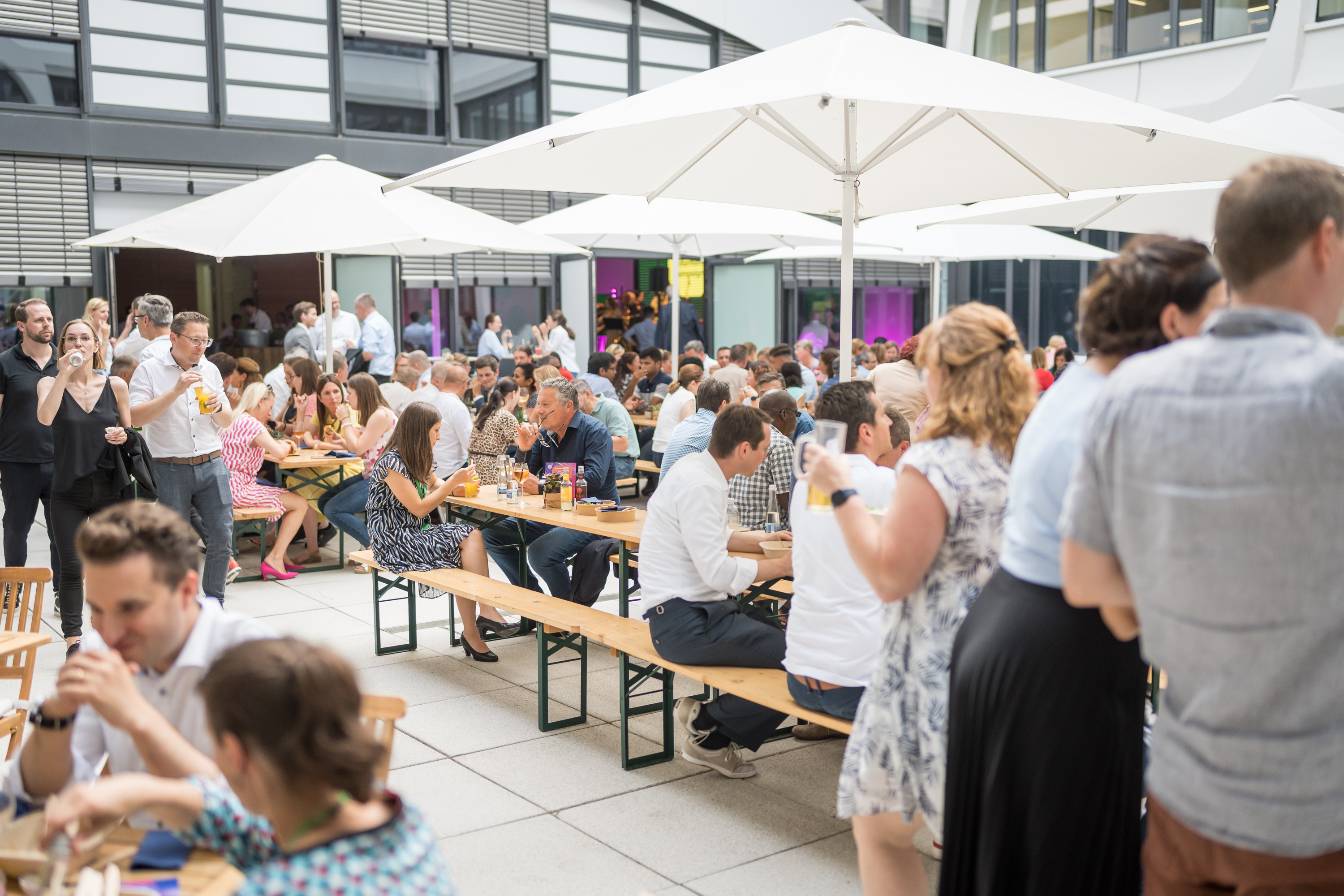 Zahlreiche Menschen sitzen und stehen an Bierzeltgarnituren unter weißen Sonnenschirmen bei einem sommerlichen Event im Innenhof eines modernen Bürogebäudes. Es wird gegessen, getrunken und sich unterhalten.