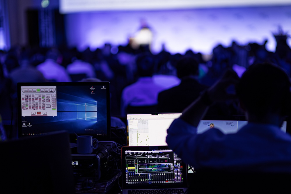 Blick von hinten auf eine große Konferenz mit blau-violetter Bühnenbeleuchtung. Im Vordergrund sind mehrere Bildschirme mit Steuerungssoftware und ein Techniker bei der Arbeit zu sehen. Die Bühne und das Publikum sind unscharf im Hintergrund.