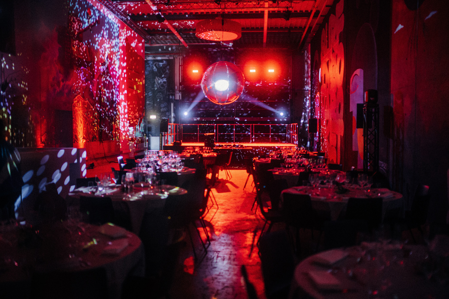Dunkler Raum mit rotem Licht, mehreren Rundtischen mit Stühlen und einer großen Discokugel
