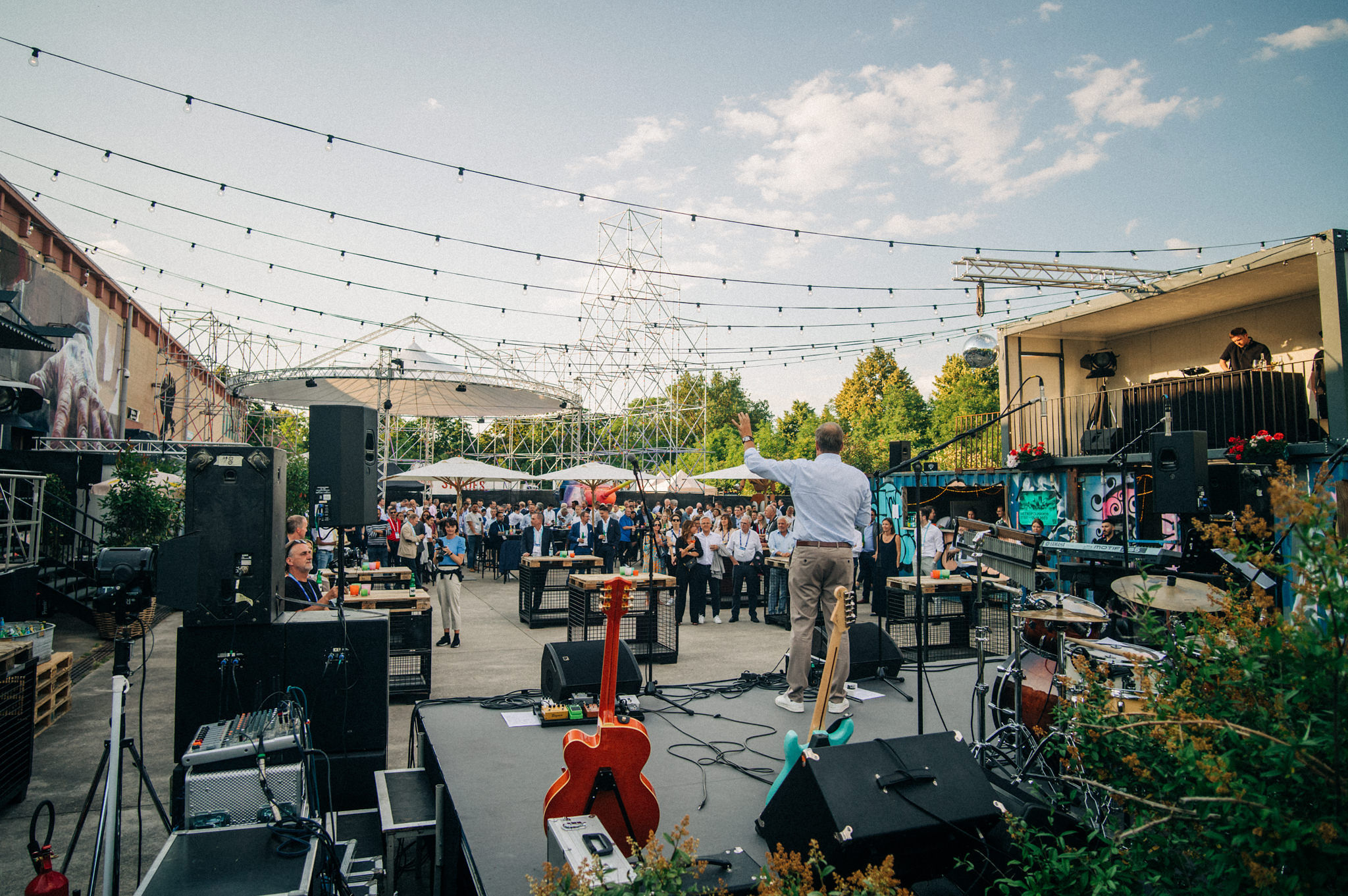 Outdoor Veranstaltung im Hellen mit einem Redner auf der Bühne und Blick auf viele Teilnehmer*innen und ein großes Gerüst mit einem Zeltdach im Hintergrund
