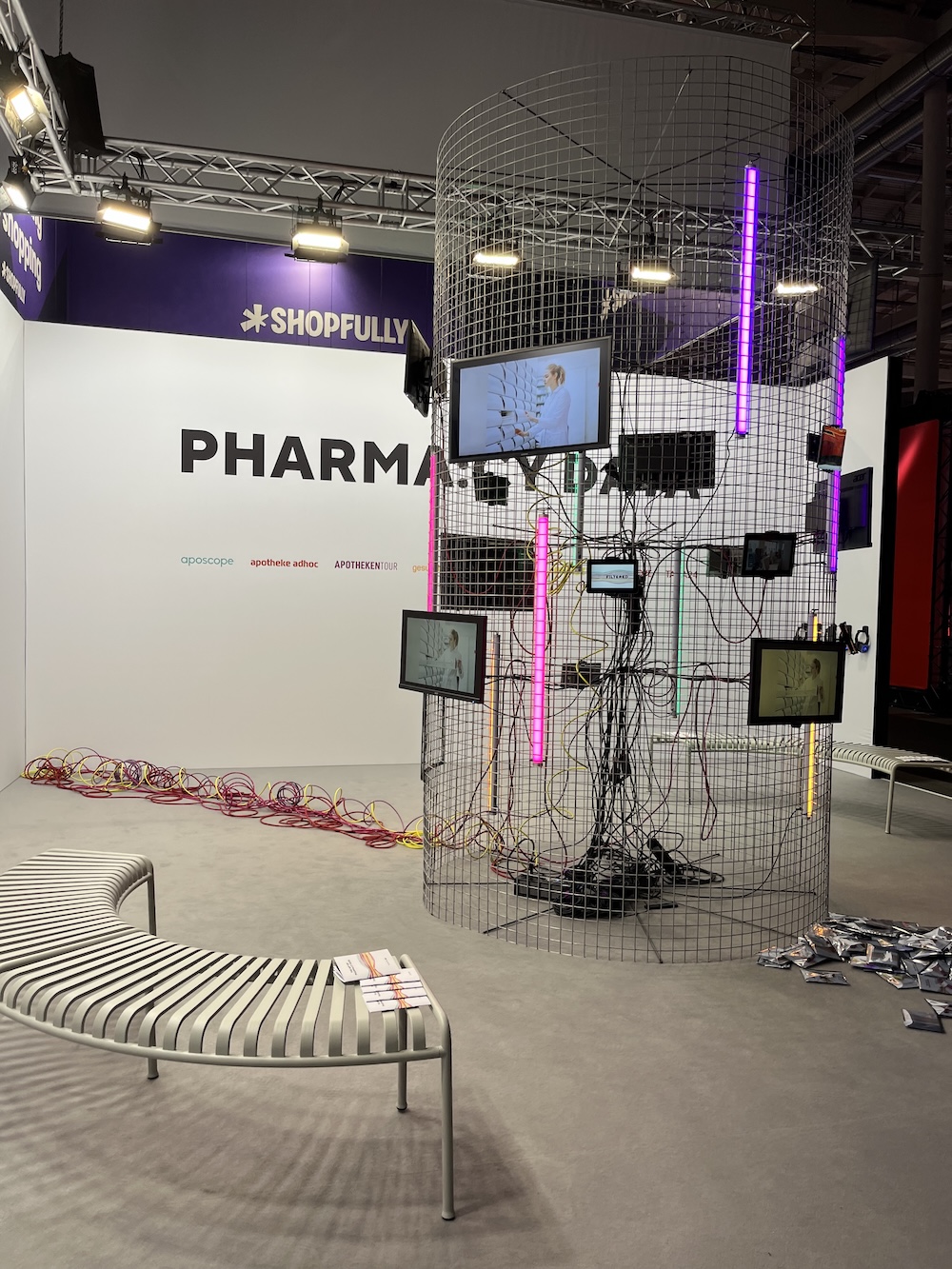 Künstlerisch gestalteter Messestand mit Gitterkonstruktion, bunten Neonröhren und Kabeln, Monitoren und Sitzbank im „Pharmacy“-Themenbereich.