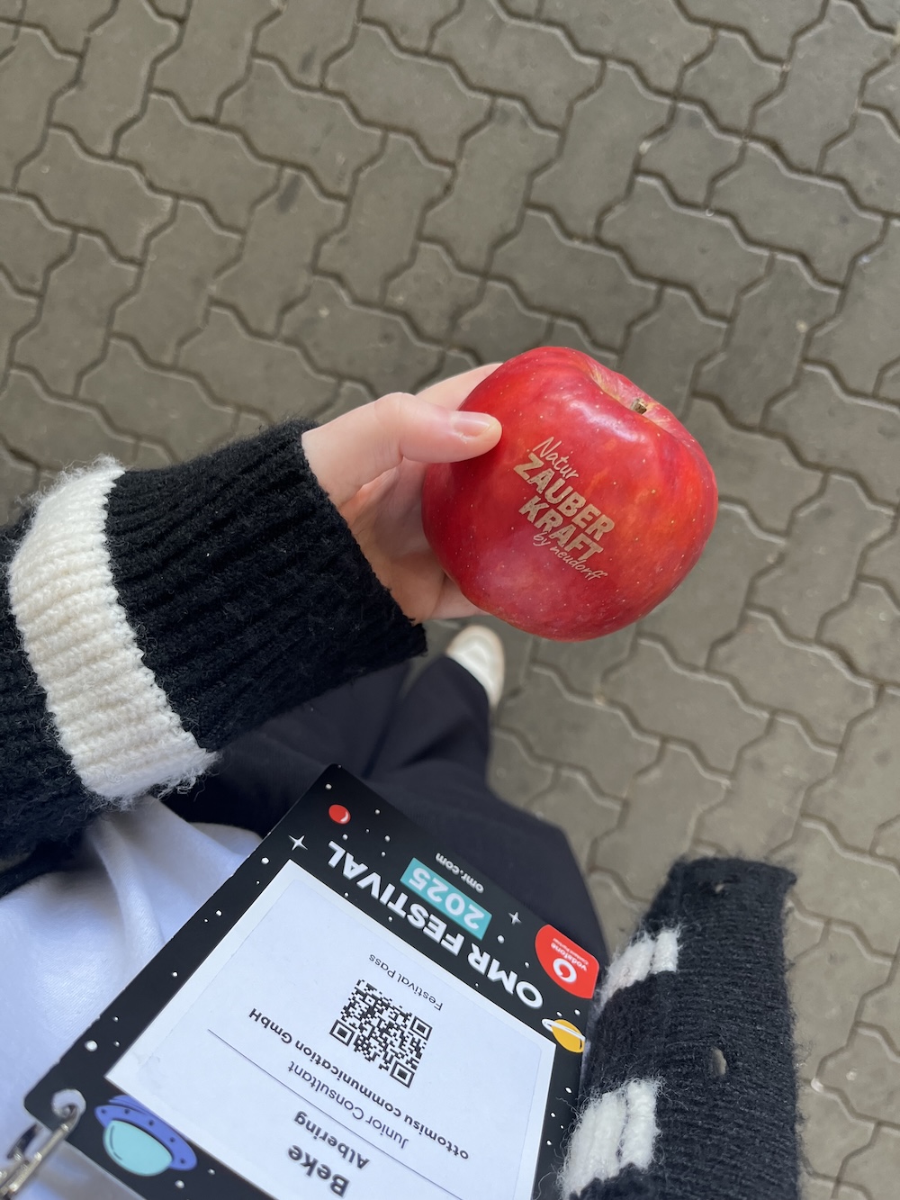 Roter Apfel mit Branding „Natur. Zauberkraft.“, in der Hand einer Besucherin mit OMR-Badge, gepflasterter Boden im Hintergrund.