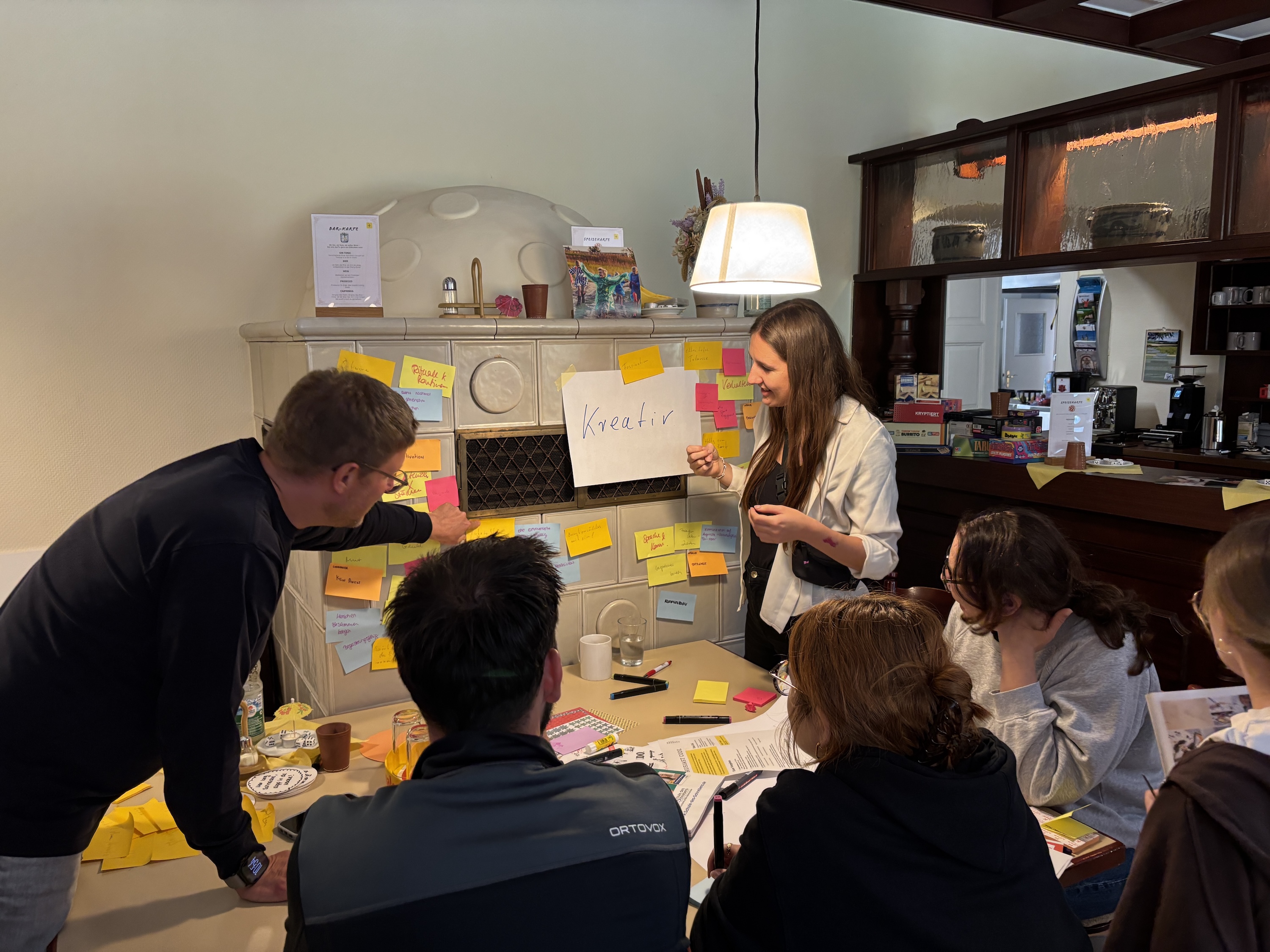 A woman moderates a creative session at a table covered with colorful sticky notes. In the background, an old tiled stove with a large sheet of paper reading “Kreativ” is visible. The group listens attentively.