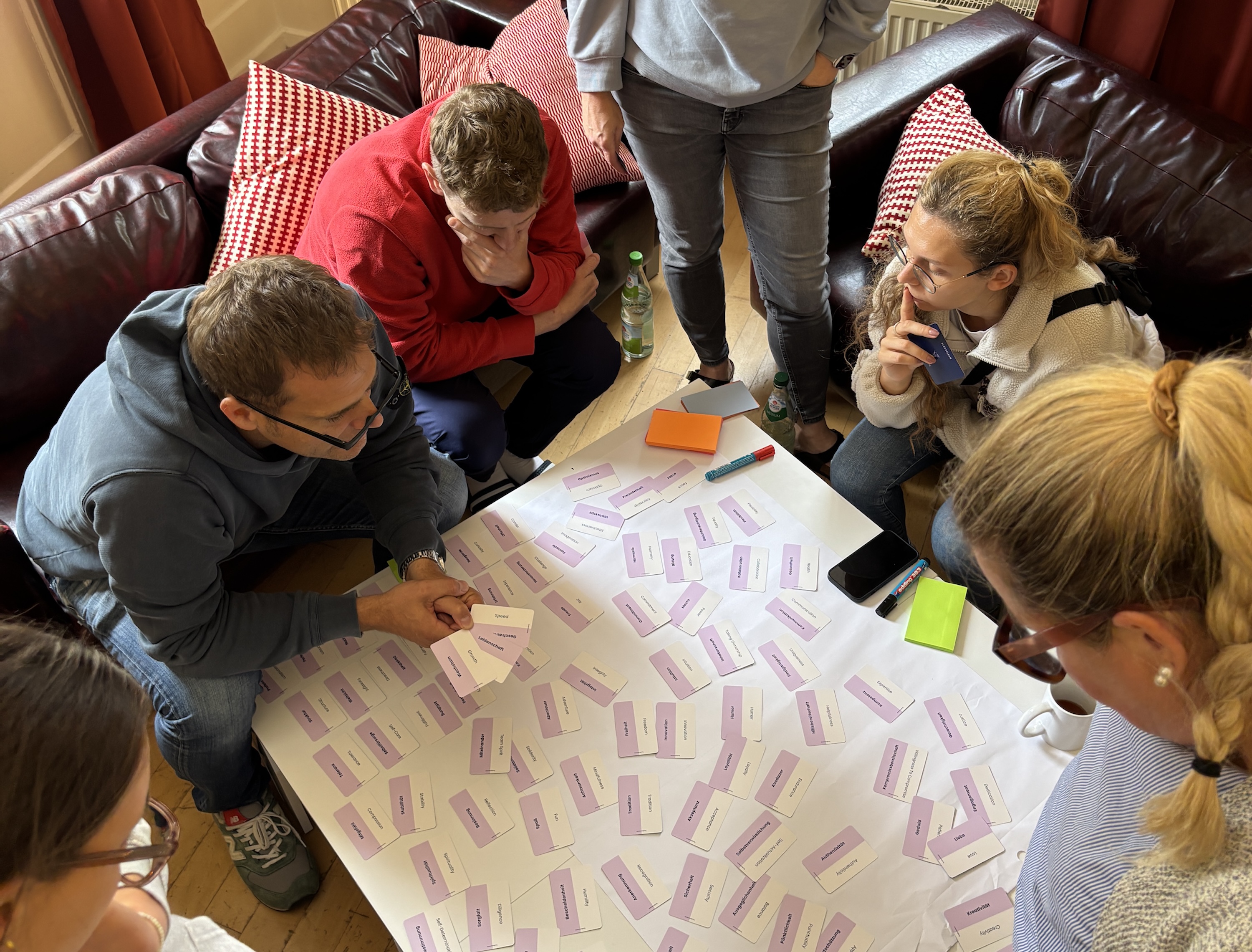 Several people sit thoughtfully around a table covered with playing cards containing words. The group is engaged in discussion. In the background, a leather couch with red cushions.