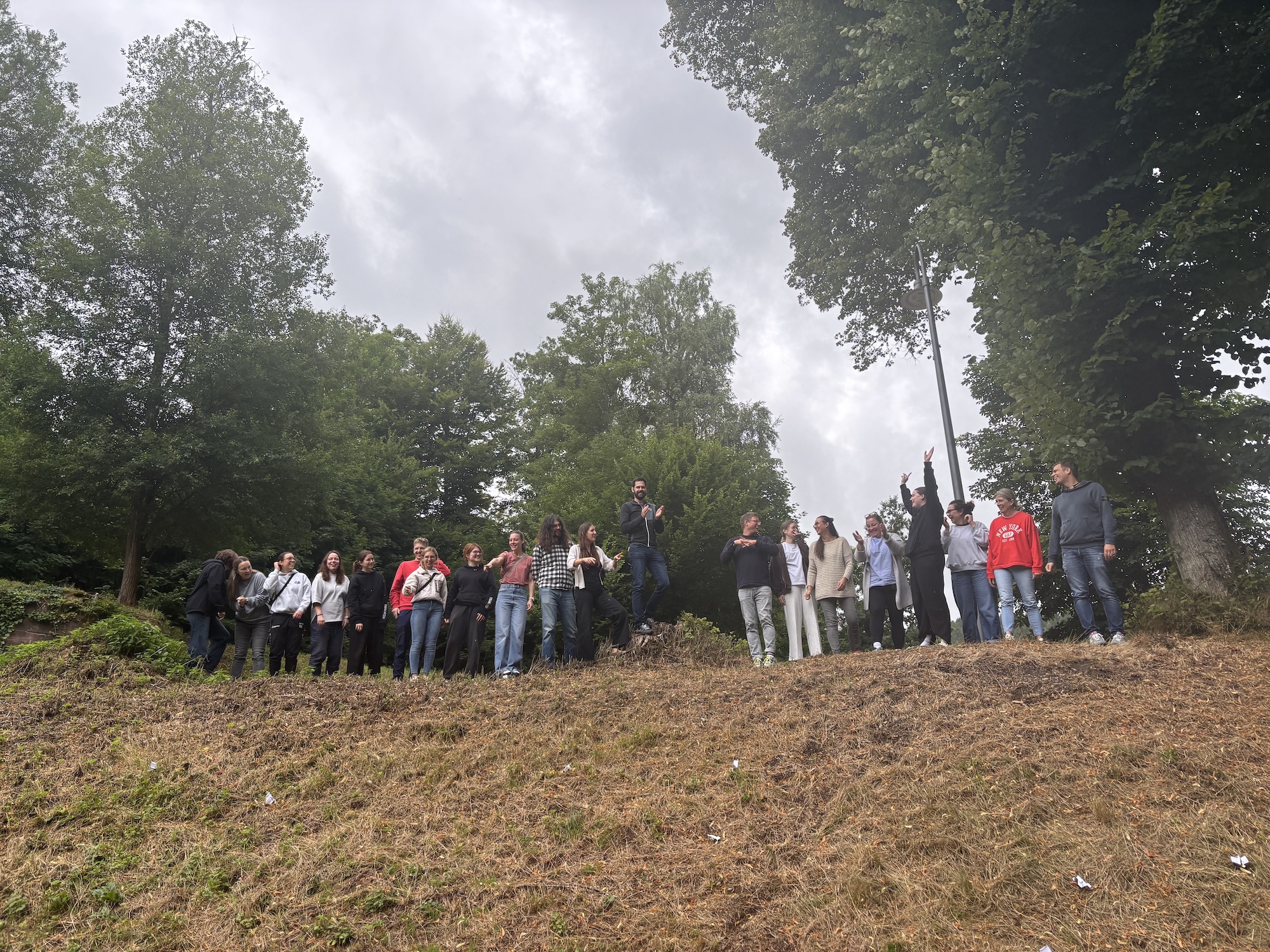 A group of people stands on a small hill in front of a wooded backdrop. Some are laughing and talking, others look ahead with curiosity. The sky is overcast.