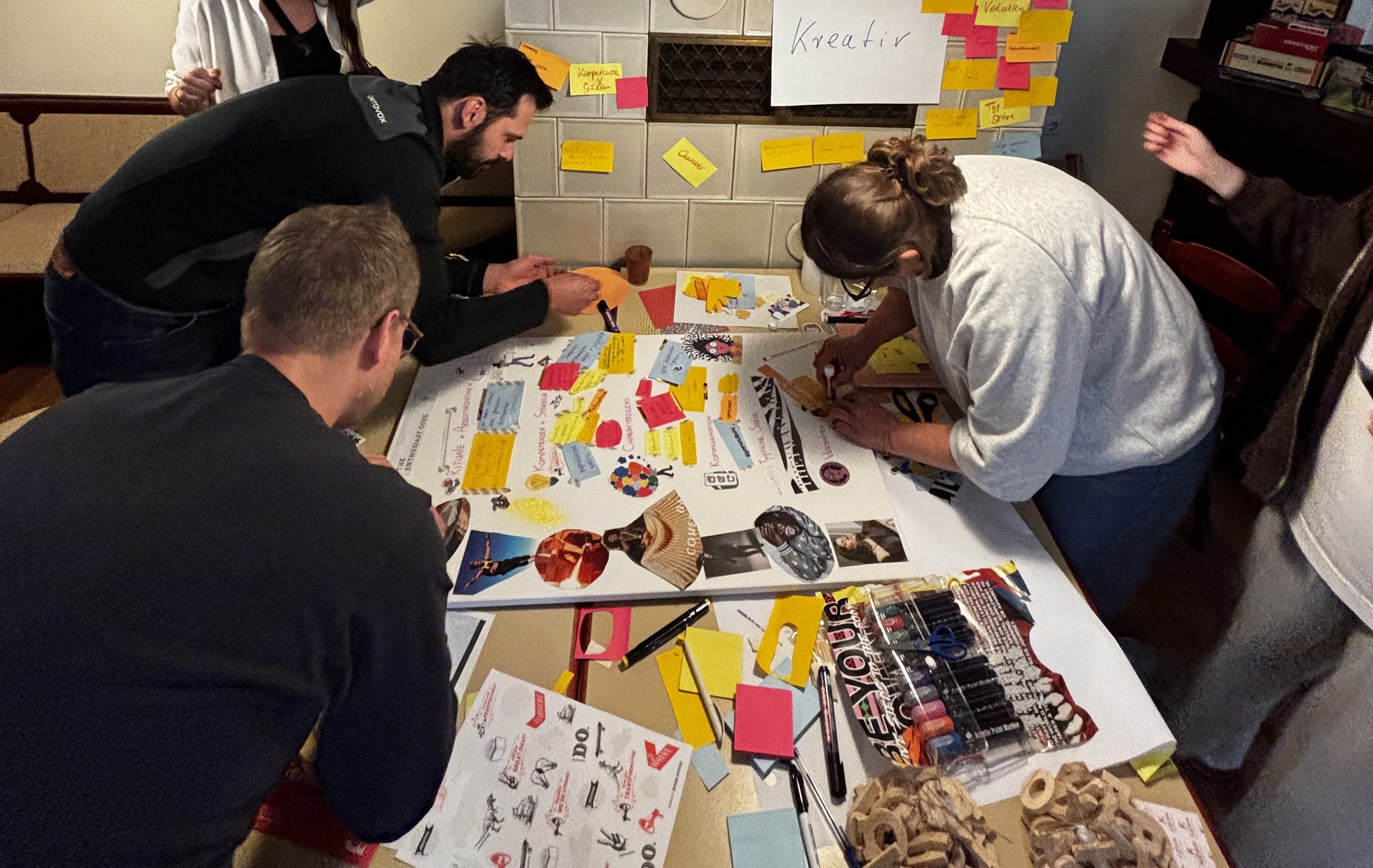 Several people work together at a large table on a collage. Scissors, pens, glue, cut-out images, and sticky notes are spread out. On the wall hangs a sign reading “Kreativ”.