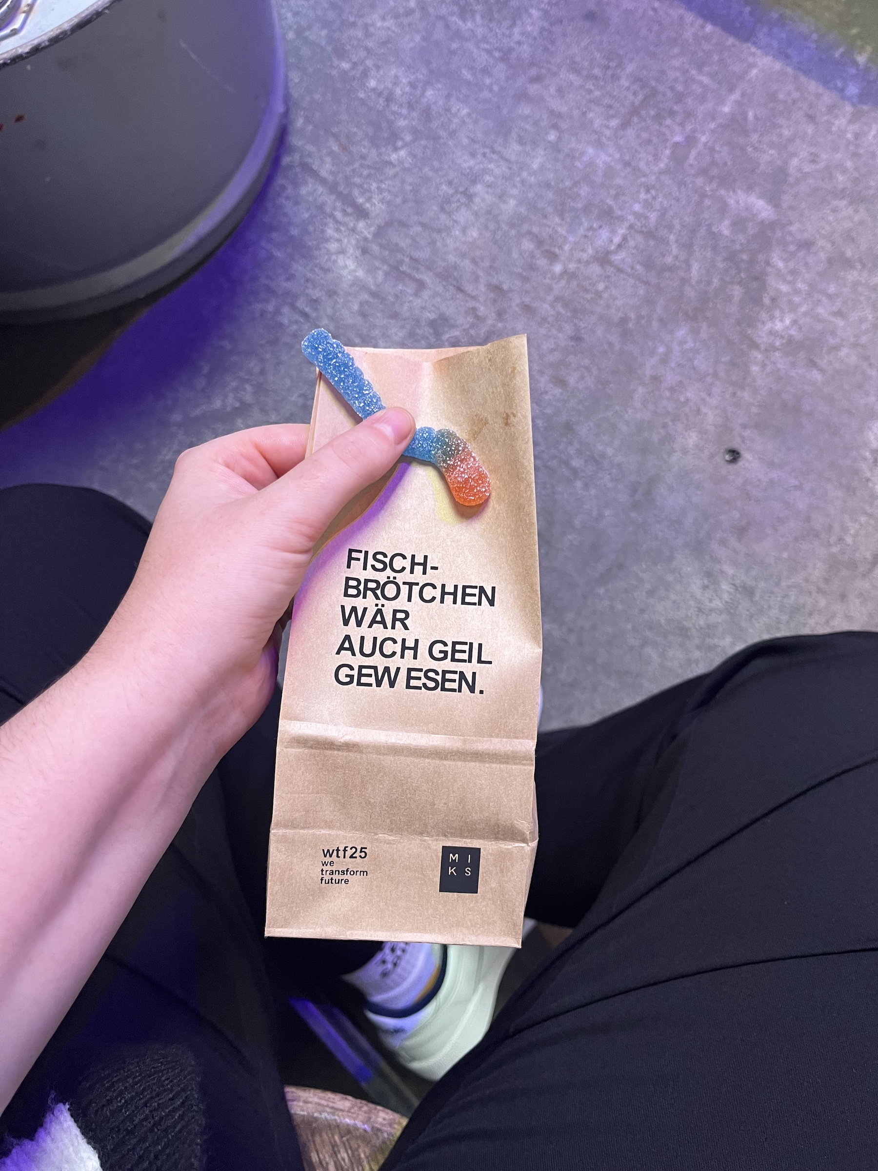 Hand hält eine braune Papiertüte mit Aufdruck „Fischbrötchen wär auch geil gew esen.“ und dem wtf25-Branding. Oben schauen zwei saure Gummifische heraus.