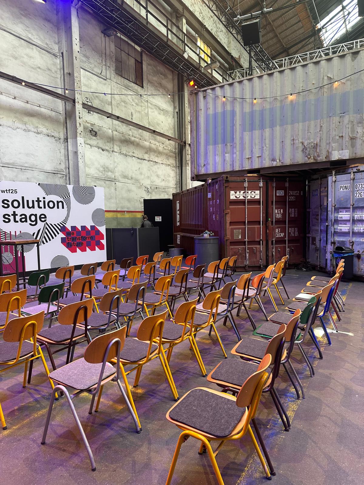 Blick auf die „solution stage“ beim wtf25-Event mit bunten Holzstühlen in Reihen, vor einem Container-Ensemble in einer industriellen Eventhalle.