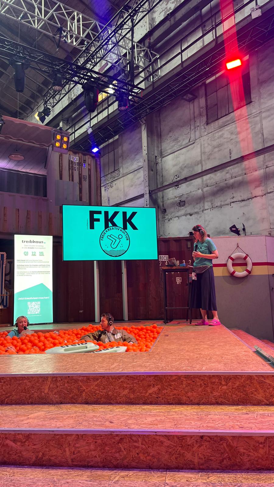 Vortragssituation auf der wtf25-Bühne mit der Präsentation „FKK – Freie Kreativ Kultur“. Zwei Personen sitzen im Bällebad, eine weitere steht am Rednerpult. Im Hintergrund ein Container-Setup und Eventtechnik.