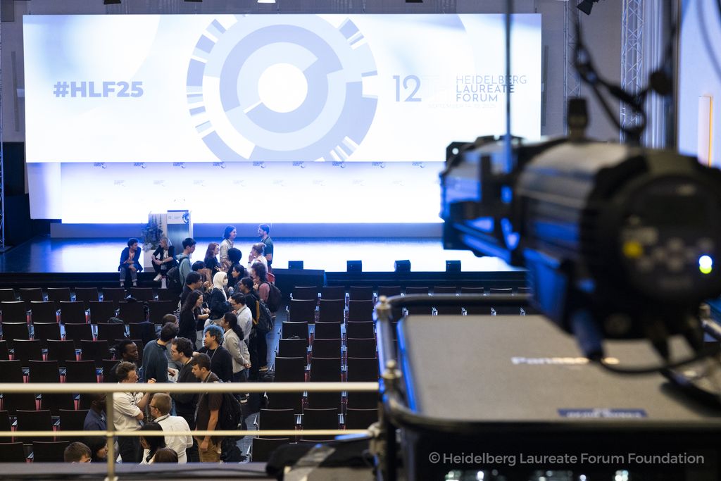 View of a conference hall with a stage and large LED screen displaying the Heidelberg Laureate Forum logo (#HLF25). In the foreground, a camera or spotlight is visible; in the hall, groups of young people are engaged in conversation between the rows of ch