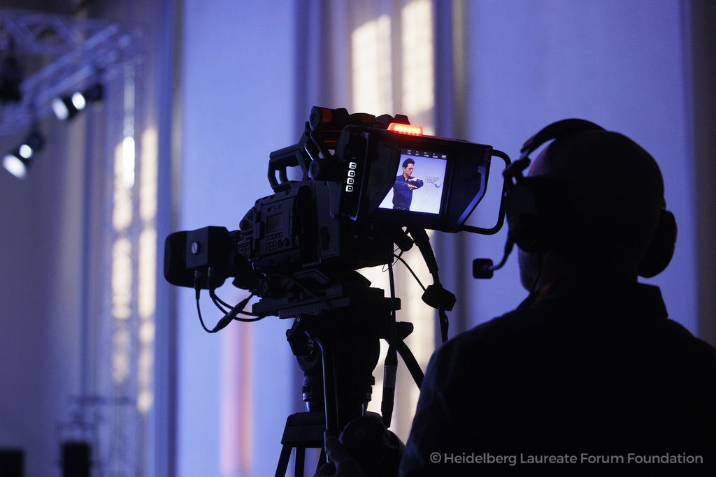 A cameraman records a presentation at the Heidelberg Laureate Forum. The stage speaker is visible on the professional camera’s display. The scene is bathed in blue lighting.