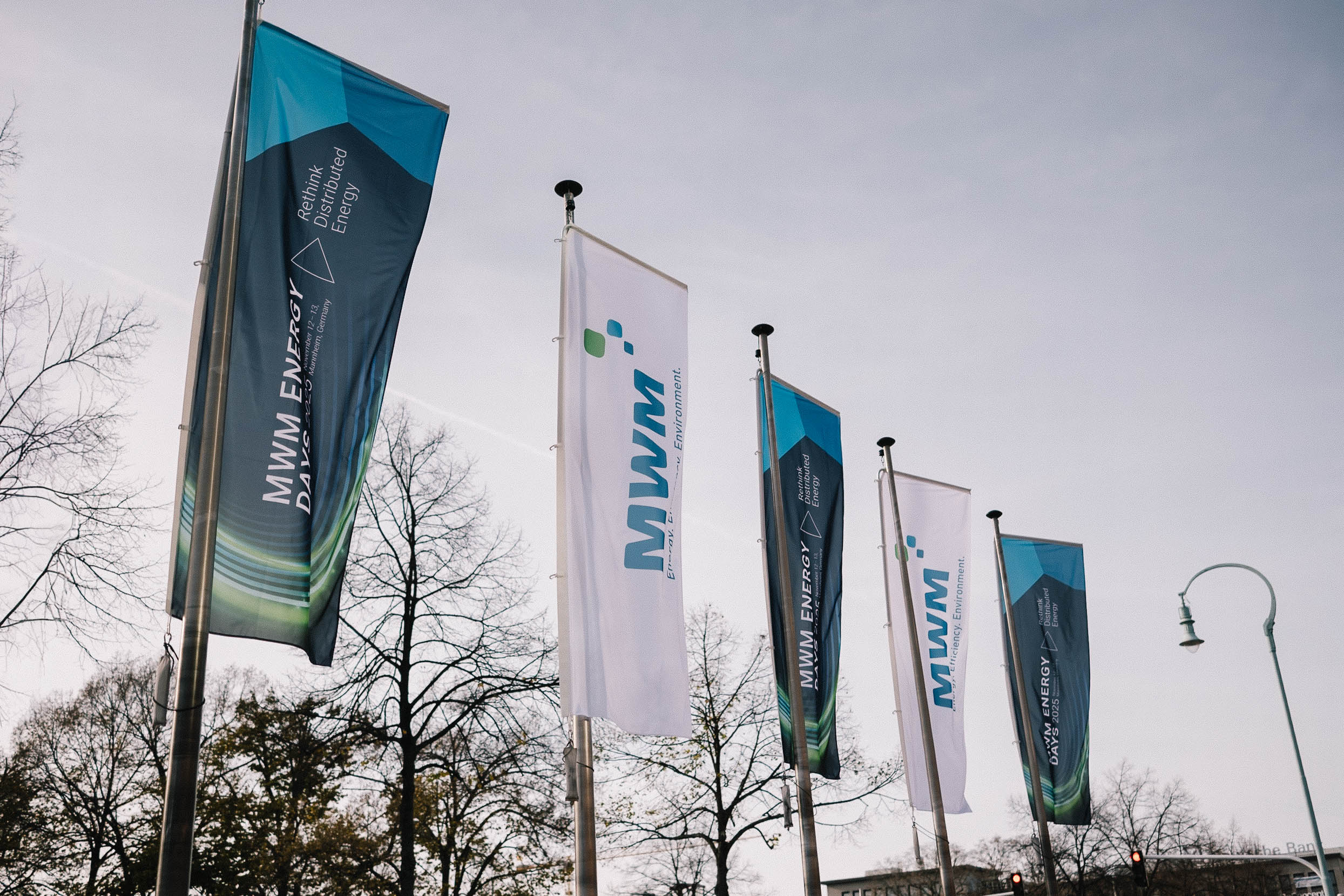 Außenaufnahme mit wehenden Event-Fahnen der MWM ENERGY DAYS 2025. Die Flaggen tragen die Slogans „Rethink Distributed Energy“ und das MWM-Logo.