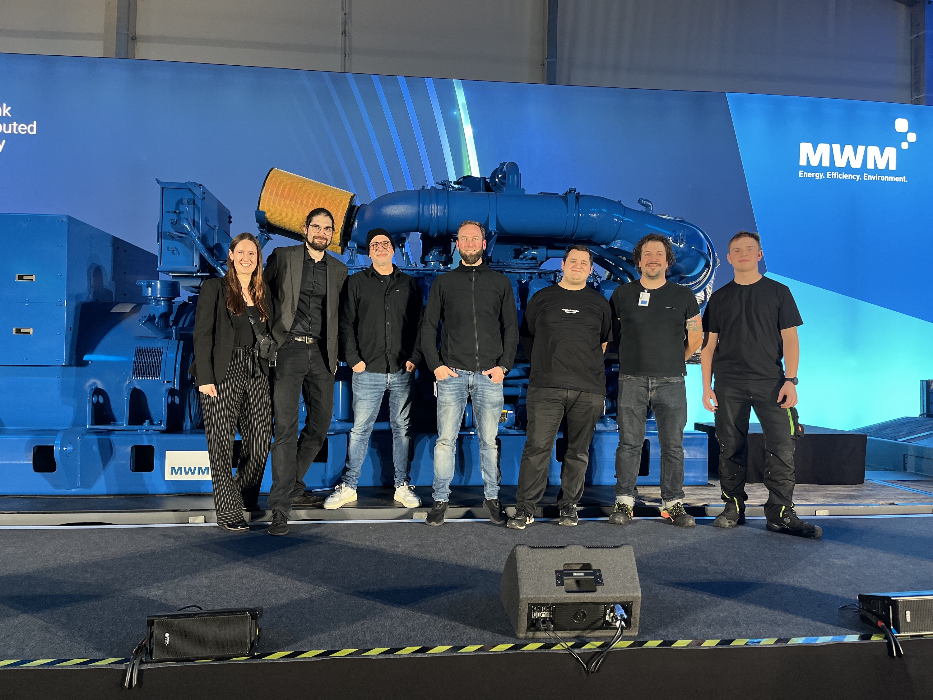 Sieben Personen stehen lächelnd auf einer Bühne vor einer großen, blauen MWM-Motoranlage. Im Hintergrund ist das Event-Branding mit den Slogans „Rethink Distributed Energy“ und „MWM – Energy. Efficiency. Environment.“ zu sehen.