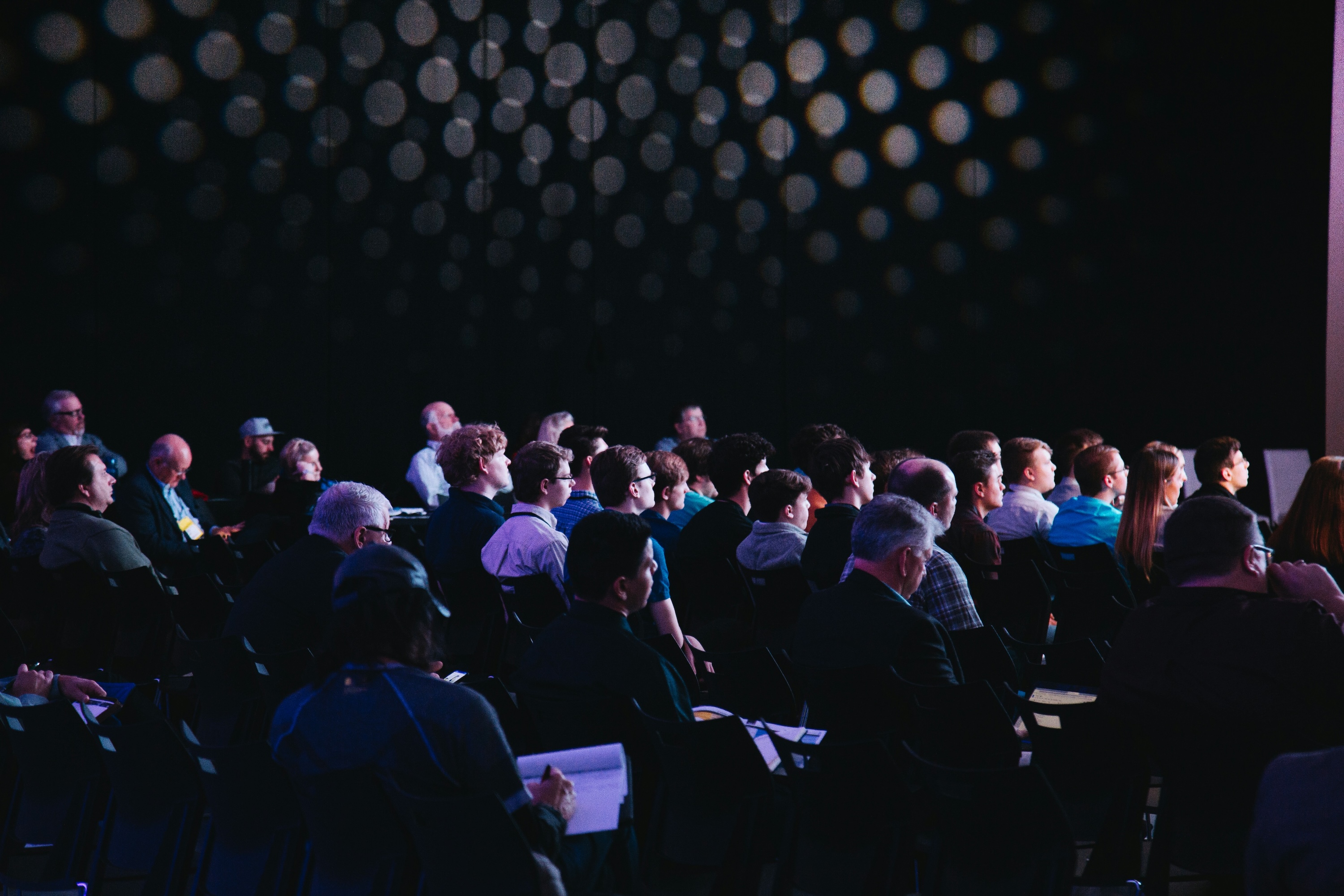 Audience seated in a dimly lit event space, attentively watching a stage out of frame, with decorative light dots visible in the background.